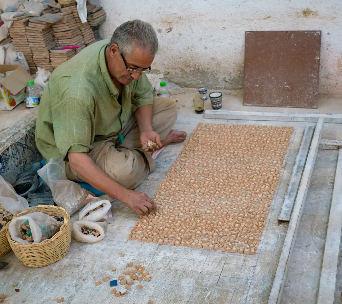 A craftsman seated on the floor places cut zellige pieces face down into a rectangular panel, with bags of sorted pieces beside him.