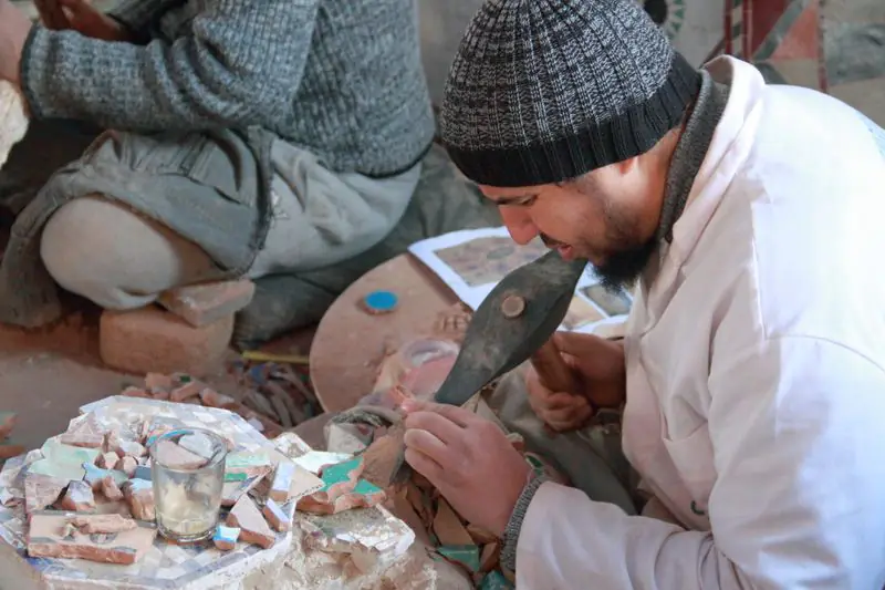 A craftsman in a white coat cuts a glazed tile into small pieces with a pointed hammer, seated among cut fragments.