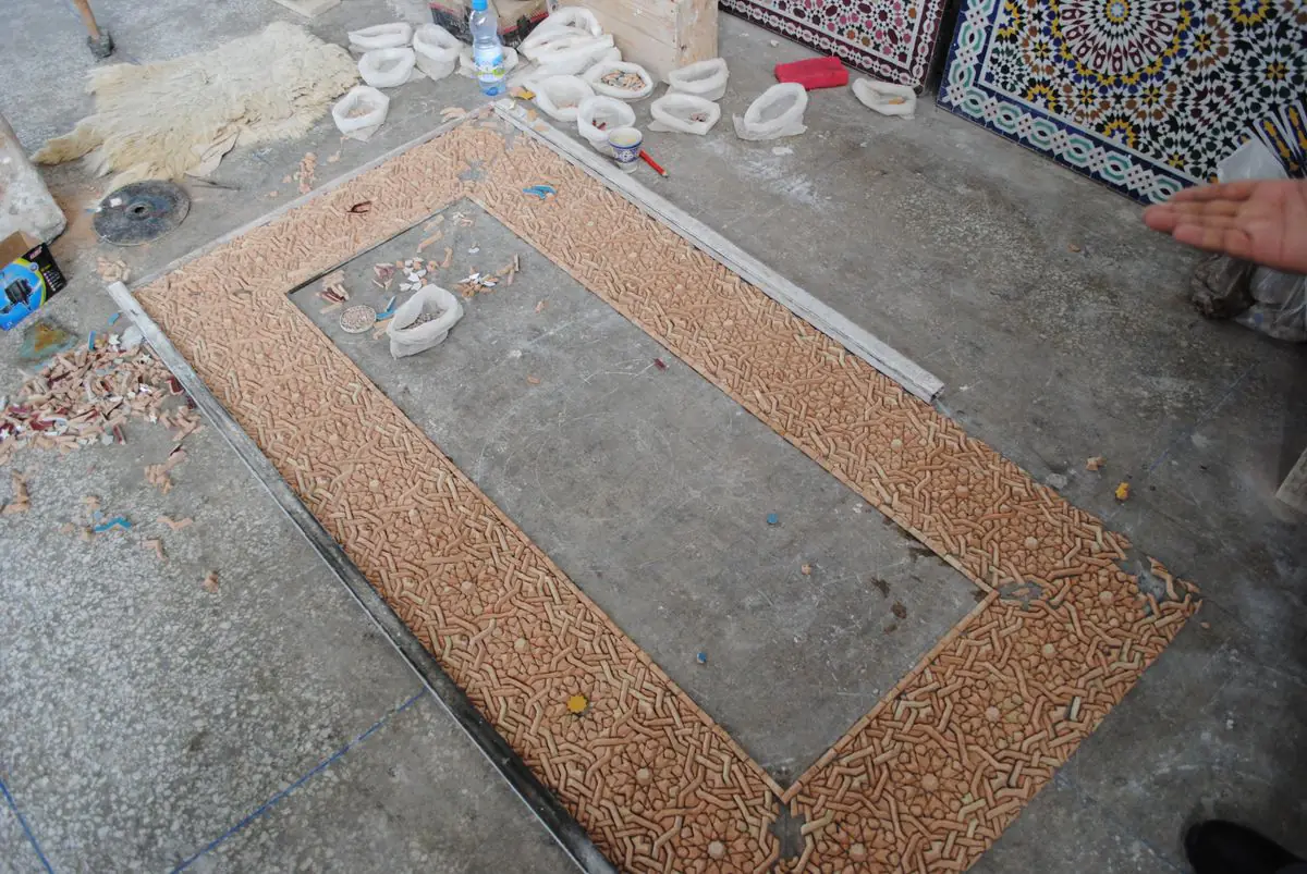 A rectangular zellige border laid face down on a workshop floor, showing the terracotta backs of the pieces, with finished panels leaning nearby.