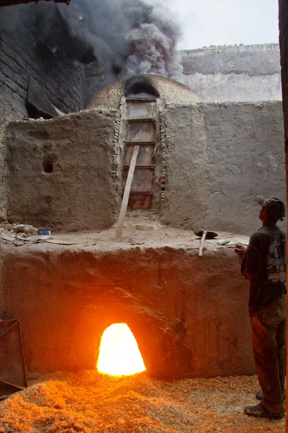 The glowing mouth of a kiln below a domed brick kiln, with a worker standing beside it.