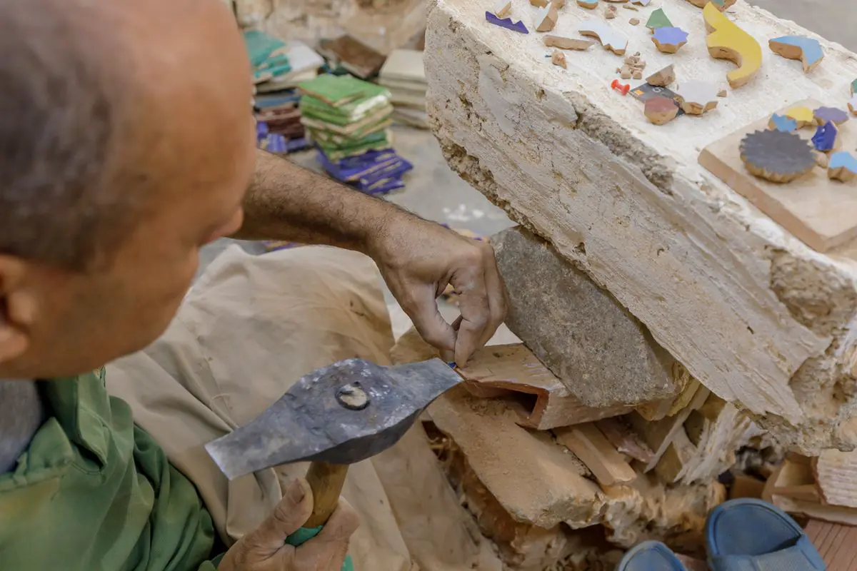 A craftsman's hand and a sharp-edged hammer trimming a glazed tile against a stone block scattered with cut pieces.