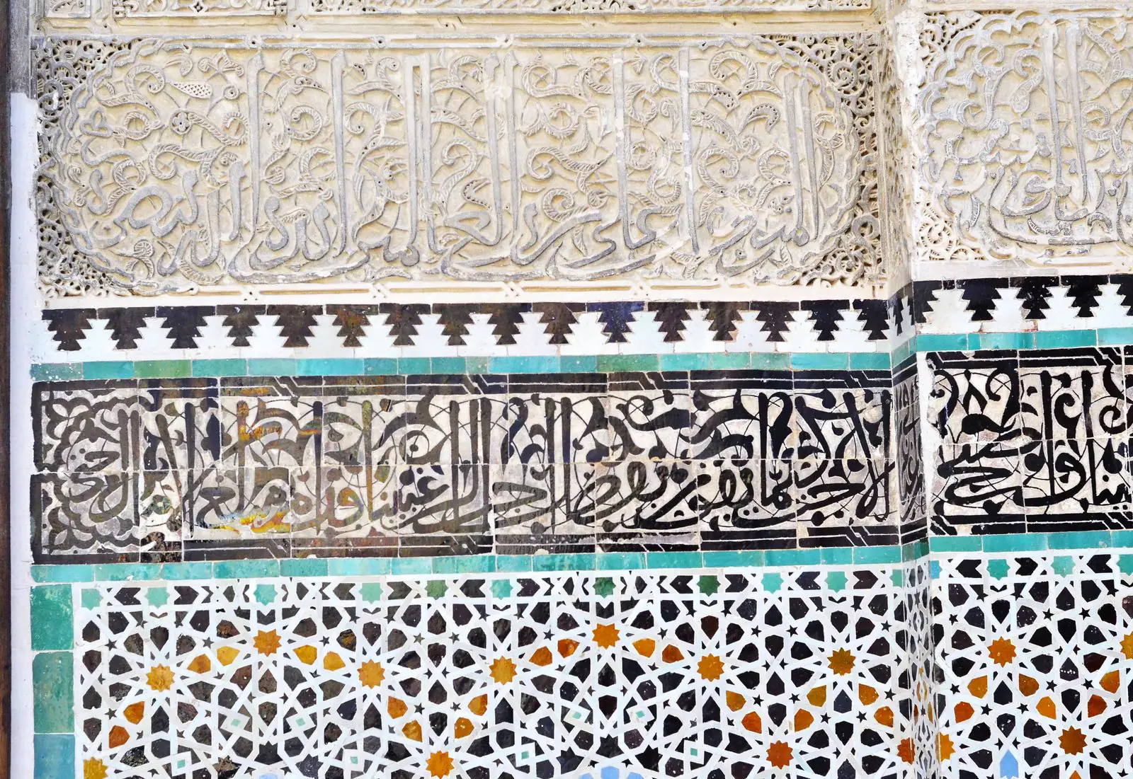 Glazed zellige in turquoise, black, white and ochre beneath a band of tile calligraphy and carved stucco at the Bou Inania Madrasa in Fès.