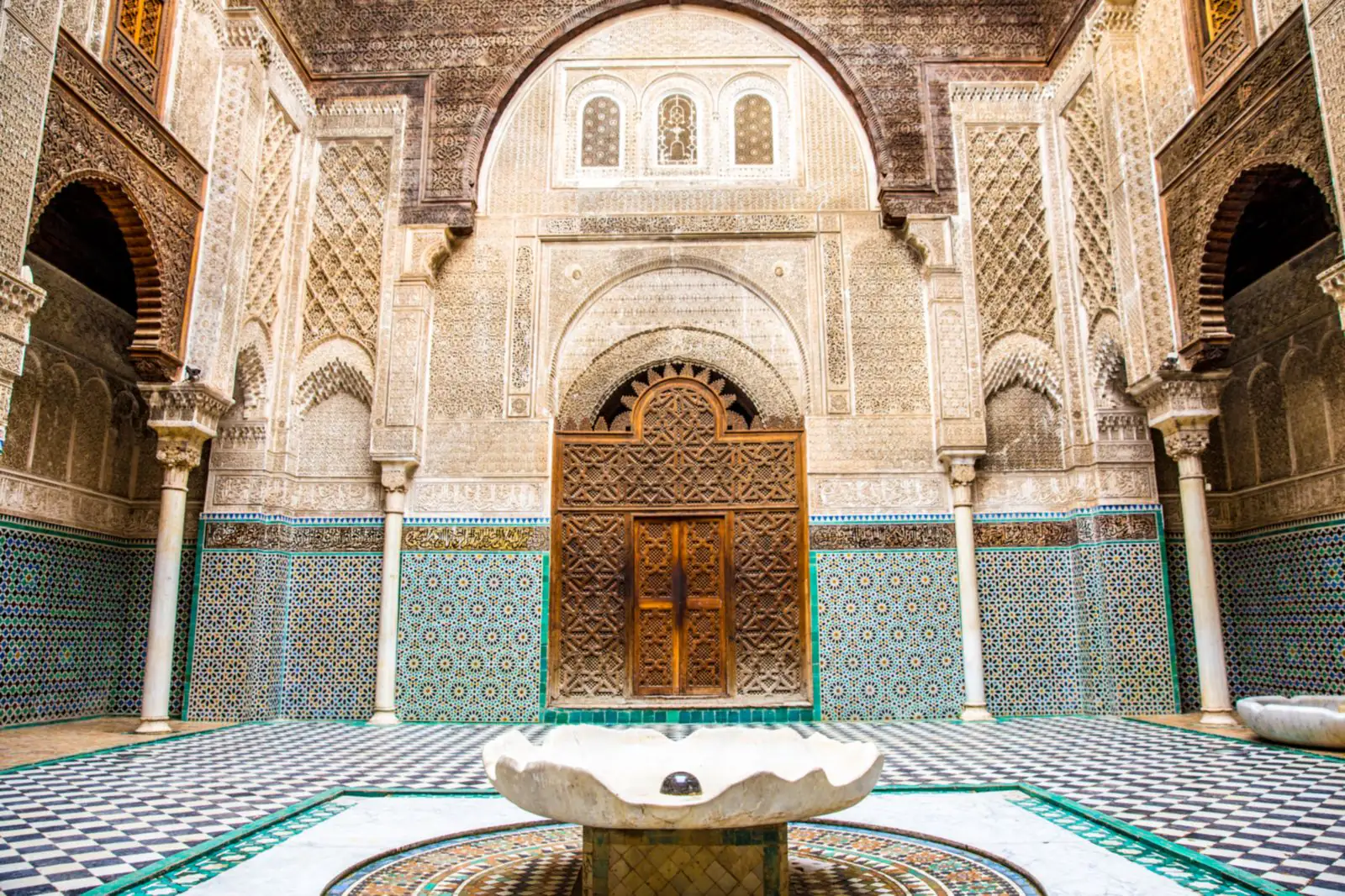 The courtyard of the Al-Attarine Madrasa: a marble basin, a zellige floor, and walls of zellige, carved stucco and cedar around a carved wooden door.