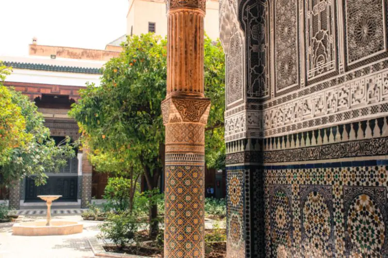 A zellige-clad column and walls of zellige and carved stucco framing a garden courtyard with orange trees and a marble fountain.