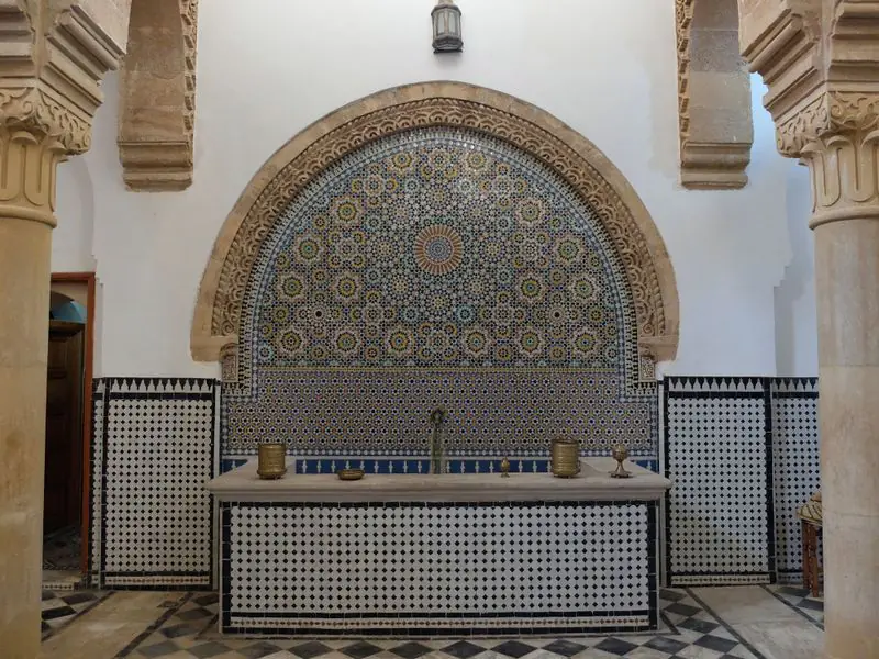 A zellige wall fountain set in a carved stone arch in a hammam reception hall, framed by stone columns.