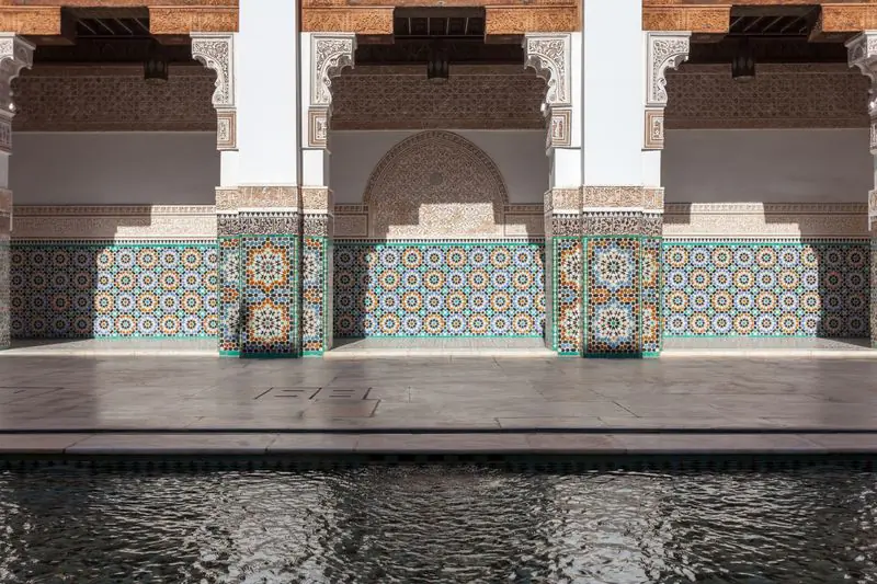 Zellige dados and carved stucco beneath a gallery, beside the still water of the courtyard pool at the Ben Youssef Madrasa.