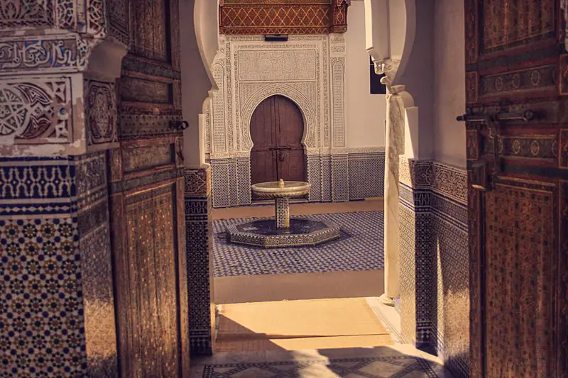 Carved wooden doors open onto a courtyard with a marble fountain, a zellige floor and zellige-clad walls.