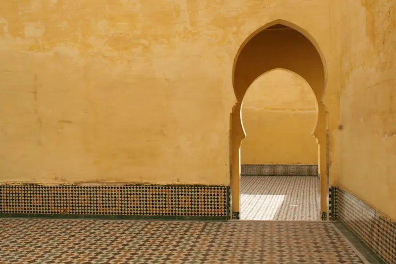 A plain ochre plastered wall above a low zellige dado, with a horseshoe arch opening onto a tiled room.